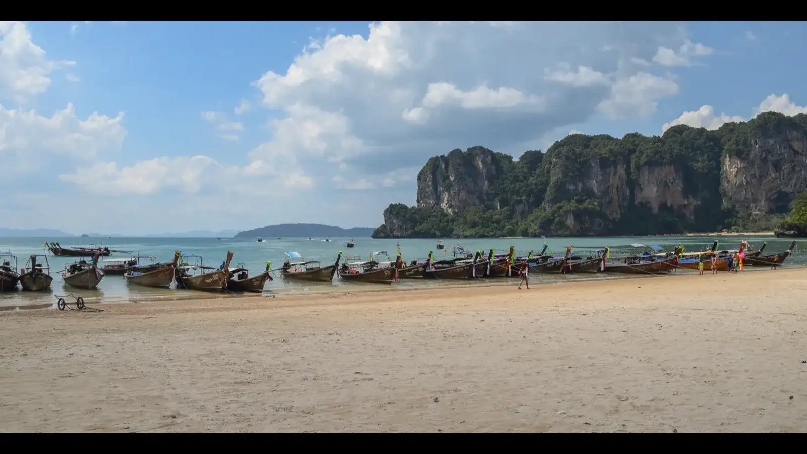 A longtail boat crossing calm turquoise water below towering limestone cliffs on a clear morning in Krabi