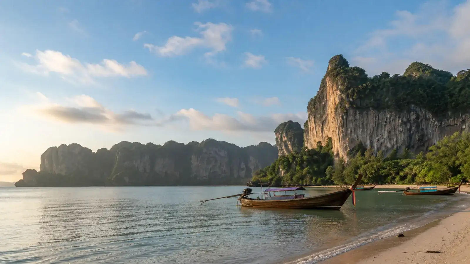 Traditional longtail boats on calm water beneath Krabi limestone cliffs during a clear early dry-season November morning