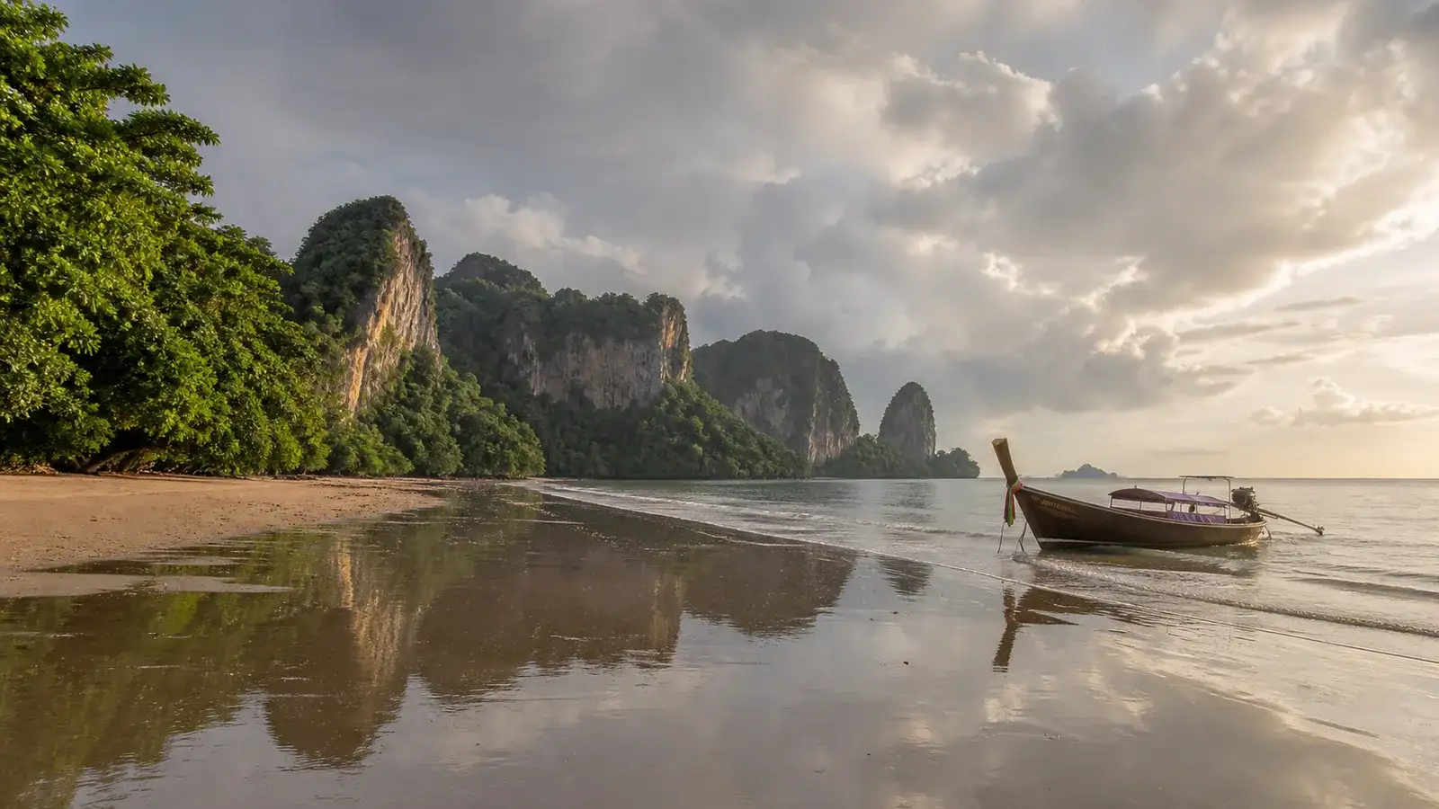 A traditional longtail boat on a reflective Krabi beach after a September rain shower, with green limestone cliffs beneath clearing clouds