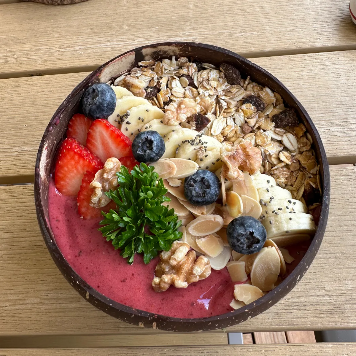 A deep-pink açaí smoothie bowl in a coconut shell topped with muesli, banana coins, sliced strawberry, blueberries, flaked almonds, walnuts and chia seeds
