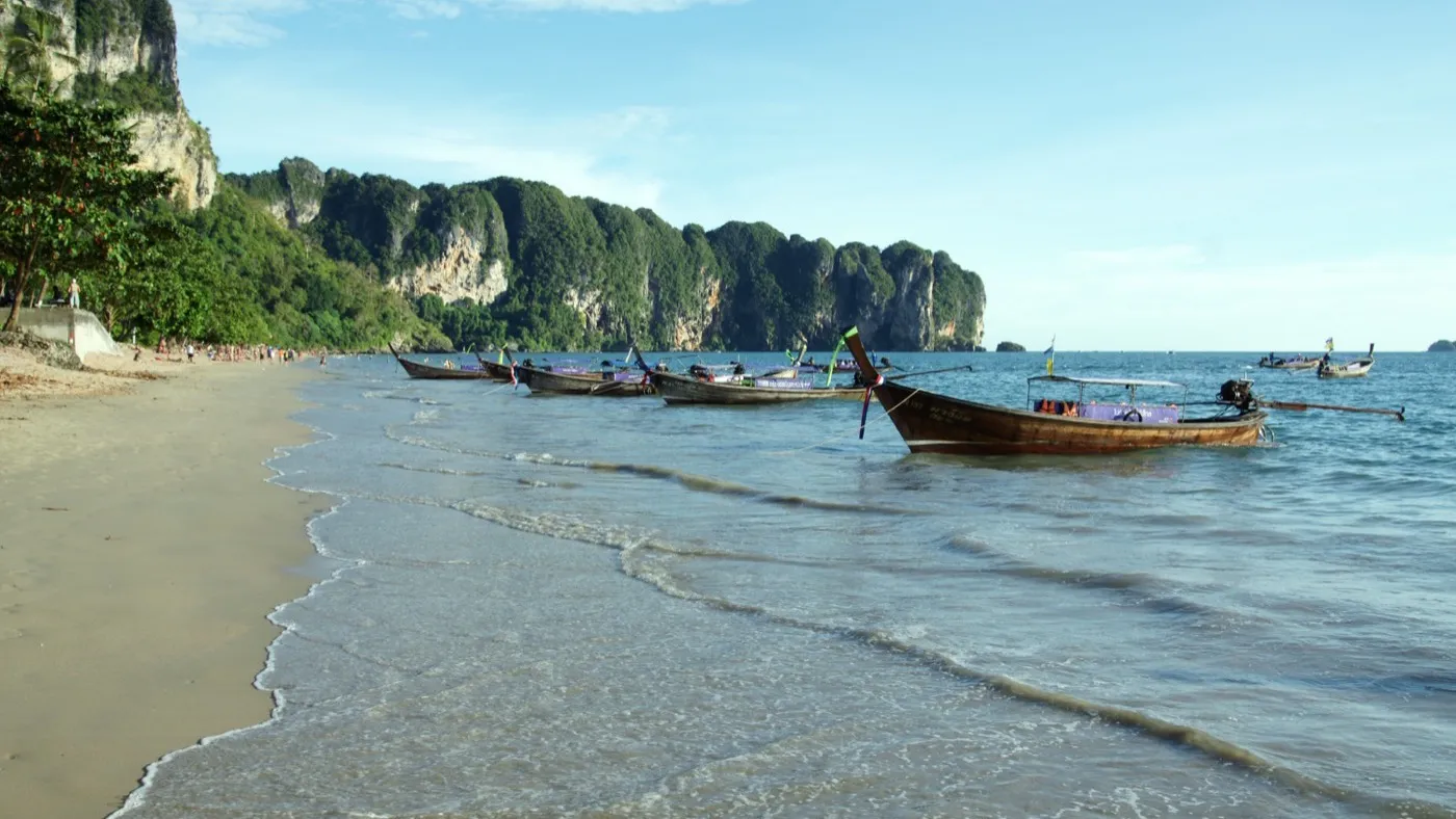 Longtail boats lined along the sand at Ao Nang beach on a sunny day, jungle-clad limestone cliffs curving away behind them