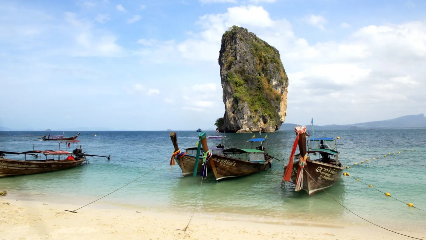Longtail boats moored off Poda Island's white beach, a tall jungle-topped limestone tower rising from turquoise water behind them