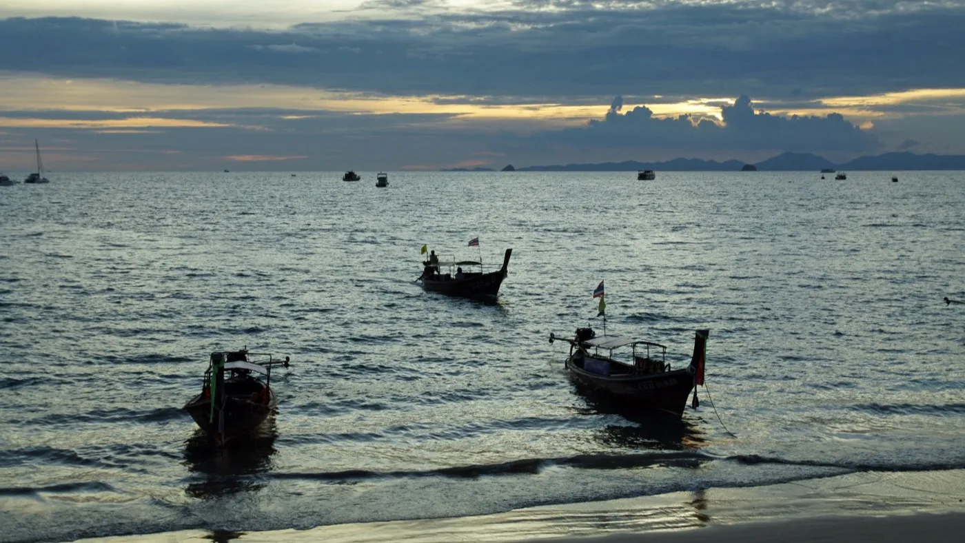 Longtail boats moored in the shallows at Ao Nang beach under an orange dusk sky, the sea calm and hills silhouetted on the horizon
