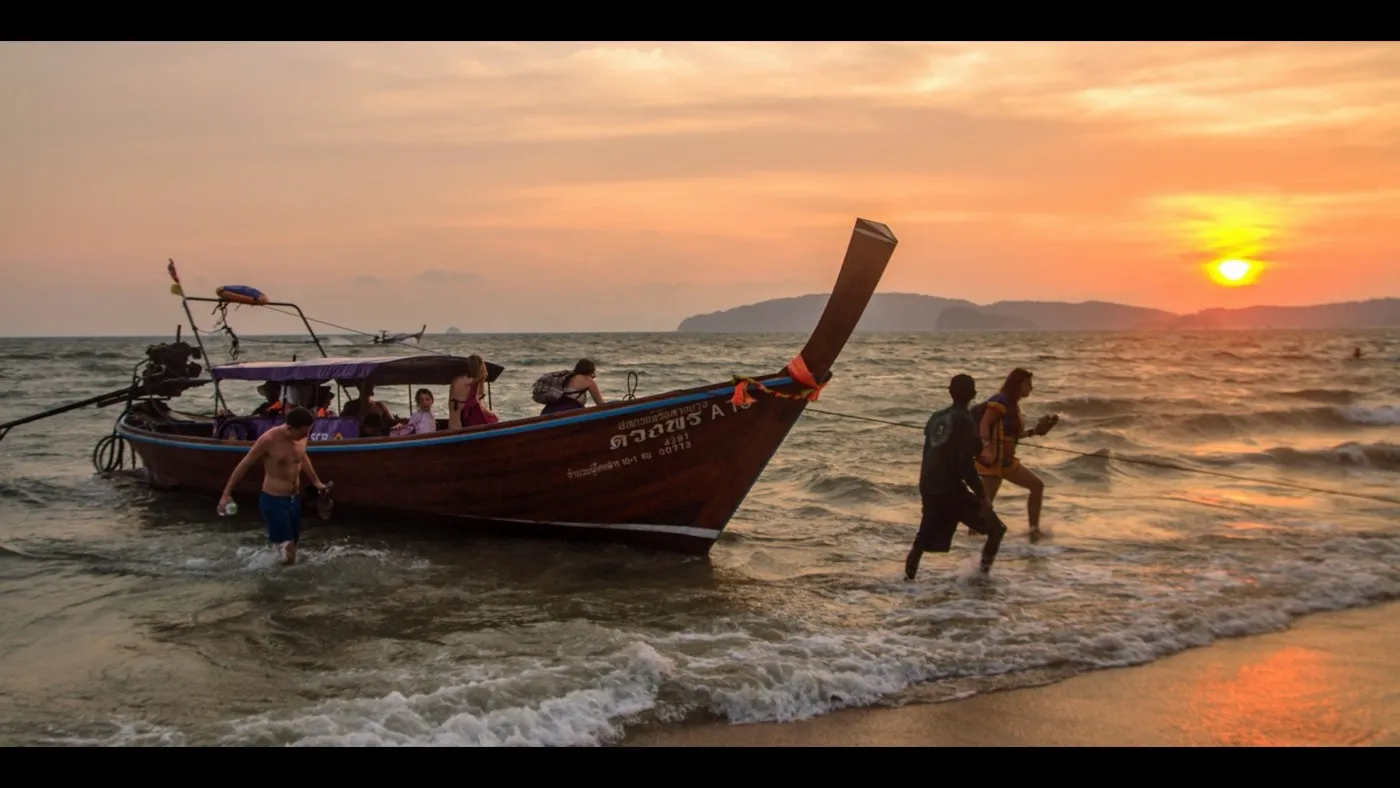 A longtail boat pulled up on Ao Nang beach at sunset, the sky glowing orange over silhouetted hills and calm sea