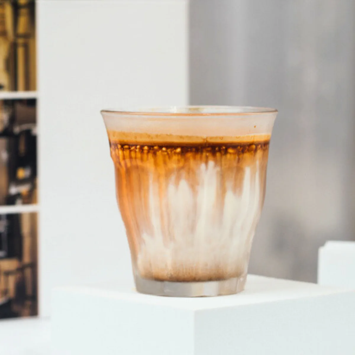 A frosted glass of dirty coffee — a dark espresso shot poured over cold milk, streaking brown through the white — on a white plinth