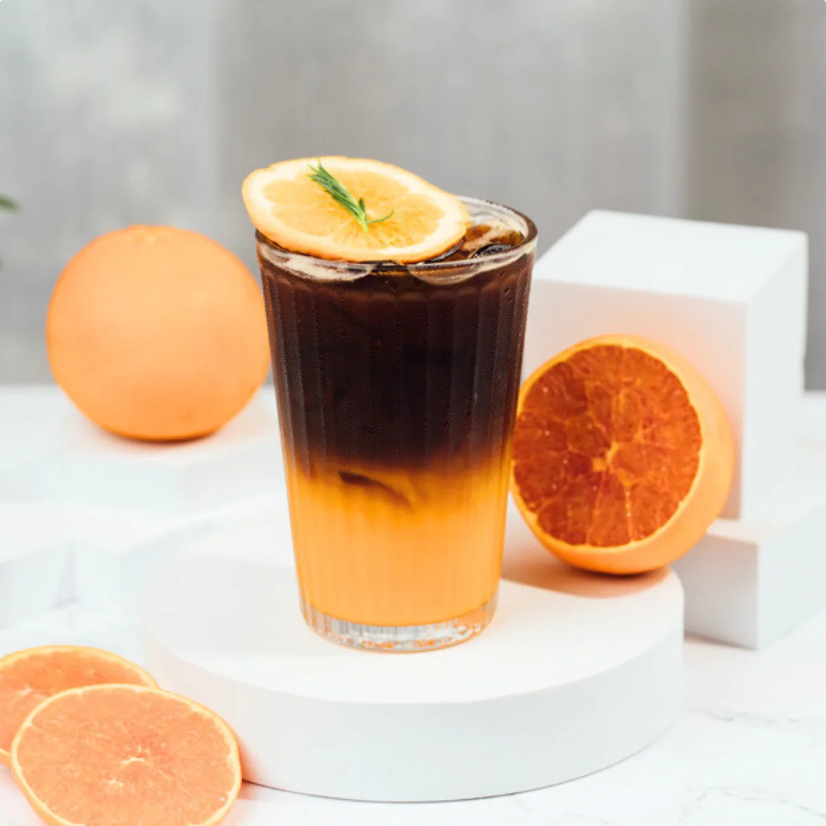 A tall glass of layered iced coffee over orange juice, garnished with a dried orange wheel and rosemary, surrounded by fresh oranges