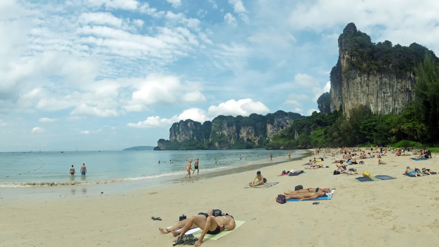 Sunbathers on the soft sand of Railay West beach beneath a sheer limestone cliff, with more karst towers along the bay