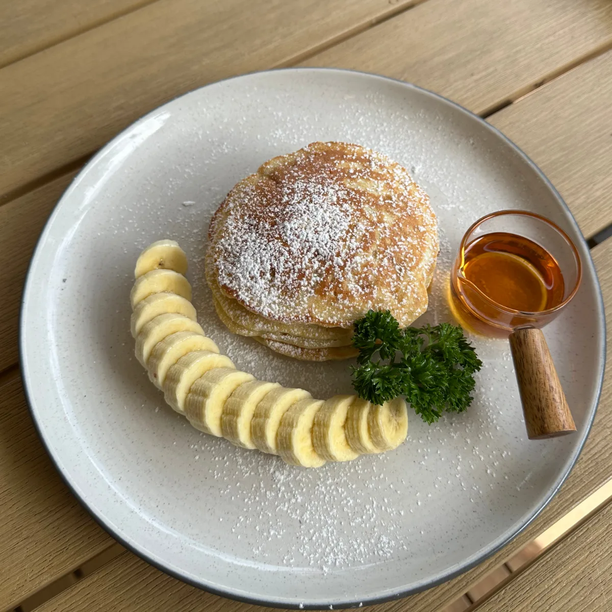 A stack of fluffy banana pancakes dusted with icing sugar, with sliced banana and a jug of syrup on a speckled plate