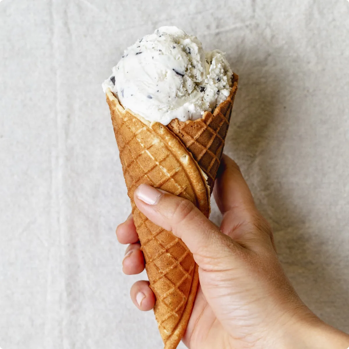 A hand holding a waffle cone of cookies-and-cream ice cream against a pale linen background