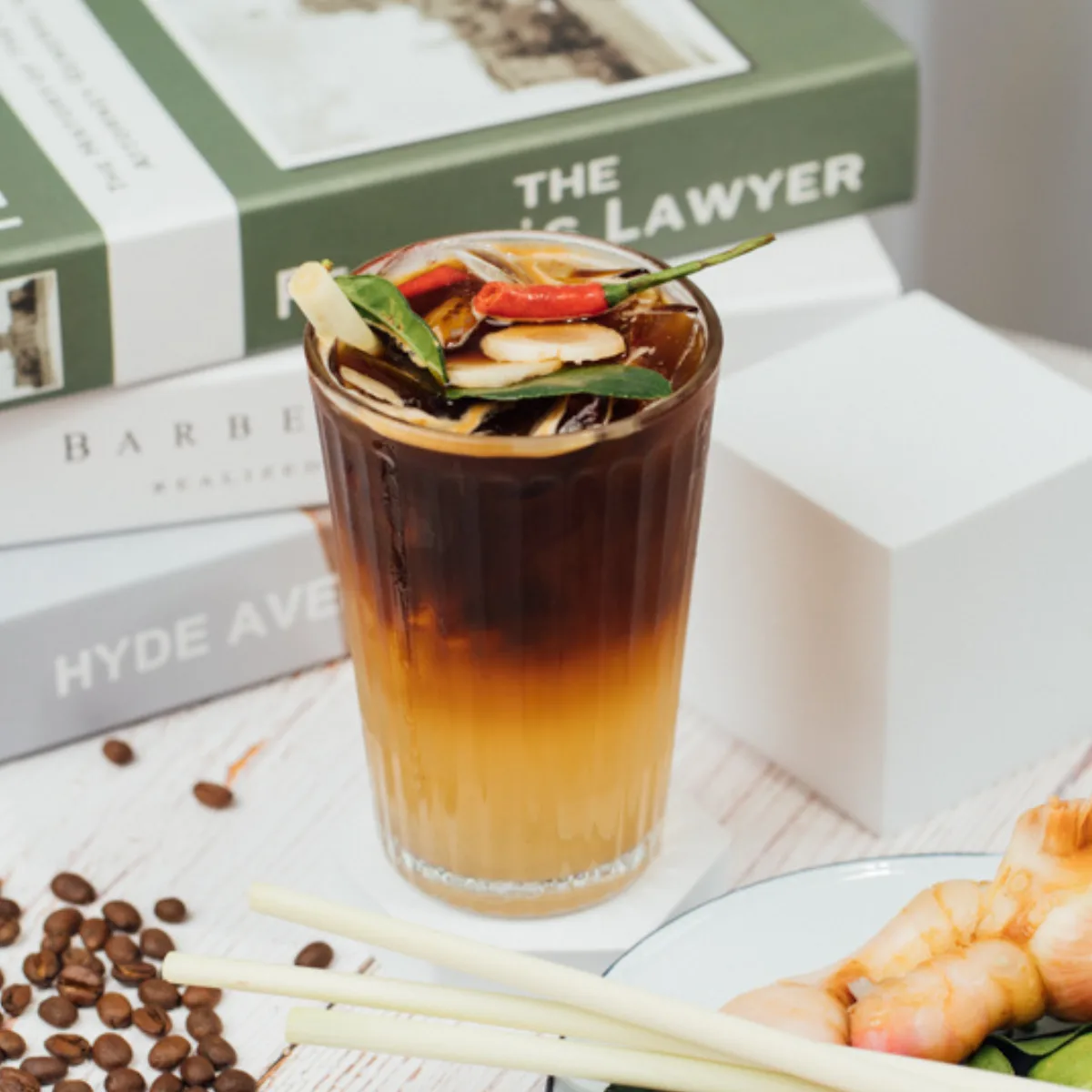 A tall glass of iced black coffee garnished with lemongrass, kaffir lime leaves and a red chili, with scattered coffee beans and prawns beside it