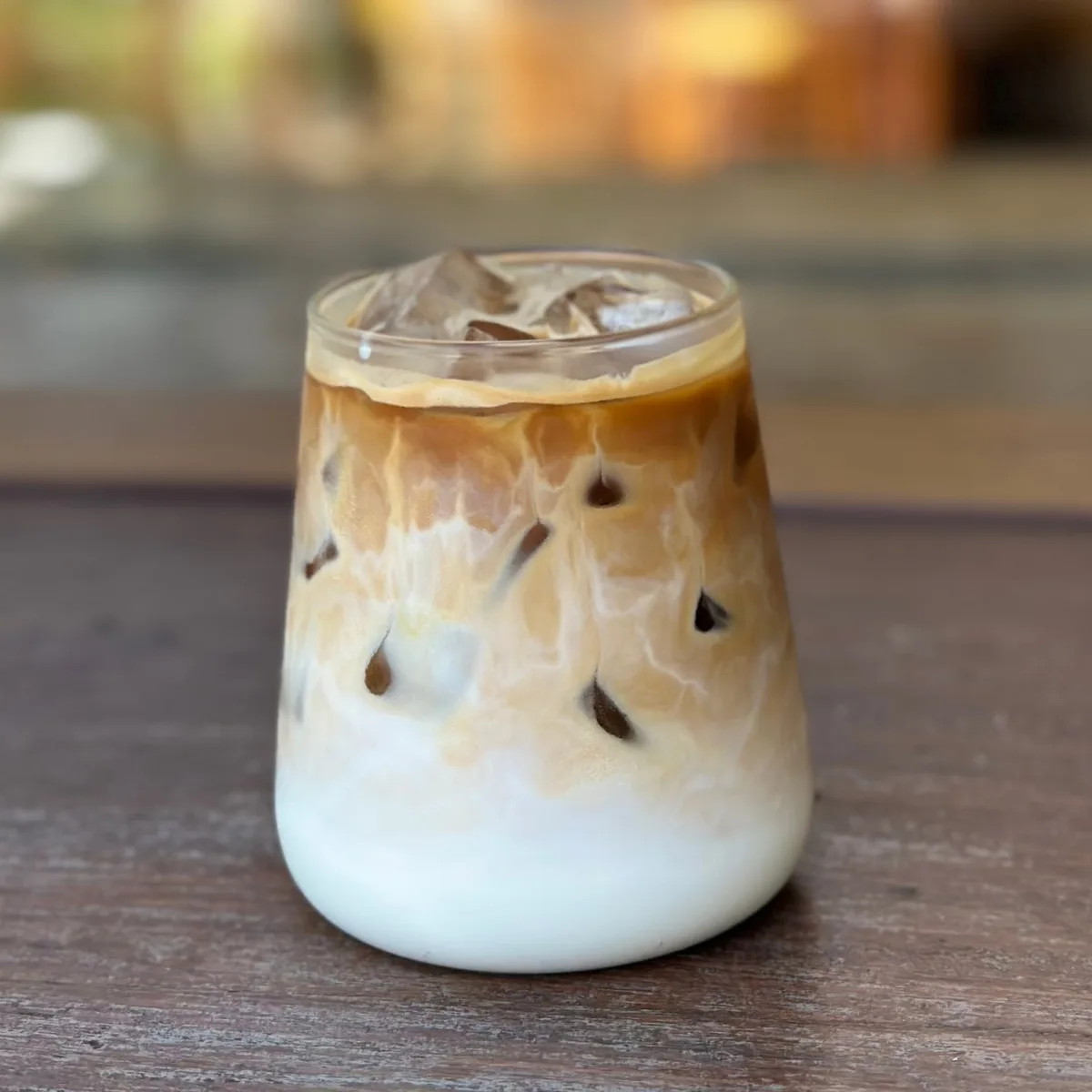 A tall iced latte in a clear glass with layered espresso and milk over ice, on a wooden table